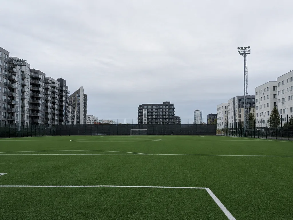 Tom konstgräsplan för fotboll omgiven av flerbostadshus och strålkastarmaster i en storstadsmiljö